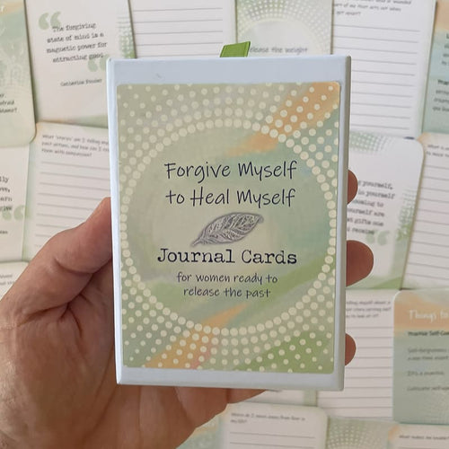A hand holds a small boxed card deck titled "Forgive Myself to Heal Myself Journal Cards, for women ready to release the past," with a green ribbon pull and a green-and-orange dotted spiral design, set against a background of scattered journal cards.