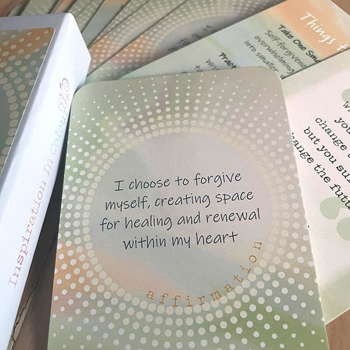 A close-up of a card reading "I choose to forgive myself, creating space for healing and renewal within my heart – affirmation," beside the closed card box labeled "Inspiration In Colour," with other cards fanned behind it.