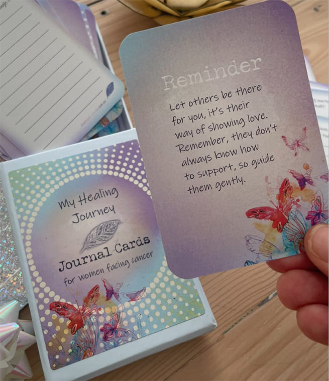 A hand holds a pastel purple journal card with a reminder message, with the card set box and another card visible on a wooden tabletop.