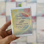 A hand holds a small boxed card deck titled "Celebrating Single Again Journal Cards, for women navigating life after divorce," with a green ribbon pull and a rainbow-toned dotted spiral design, set against a blurred background of scattered journal cards.