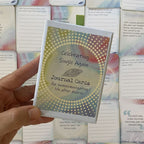 A hand holds the closed "Celebrating Single Again Journal Cards" box against a background of scattered cards showing advice text such as "Build a Strong Support System" and a quote reading "You don't have to see the whole staircase, just take the first step.