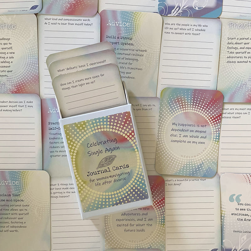 The open card box with a stack of cards inside showing prompts like "What 'defeats' have I experienced?", surrounded by fanned-out advice and affirmation cards, including one reading "My happiness is not dependent on anyone else; I am whole and complete on my own.