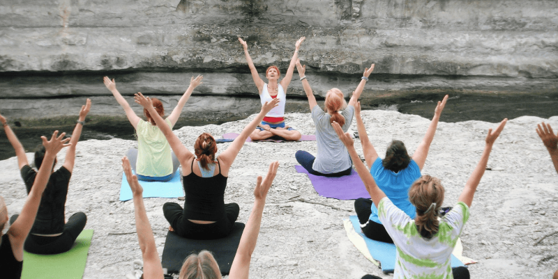 A group of people practicing yoga outdoors on a rocky cliffside, facing a cliff face with their arms raised overhead. They are seated on colorful yoga mats, including green, blue, purple, and black mats.