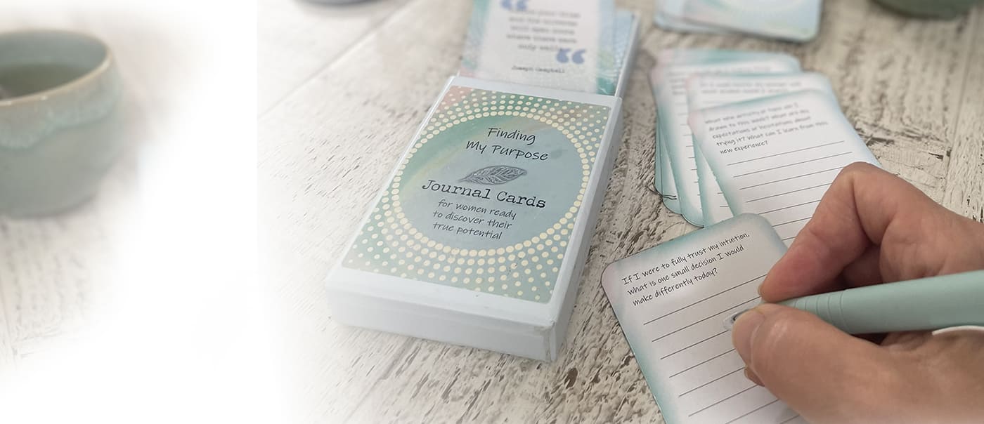 A person's hand holding a mint green pen and writing on a small lined card, with a deck of journaling cards labeled 'Journaling Cards' on a white wooden surface.